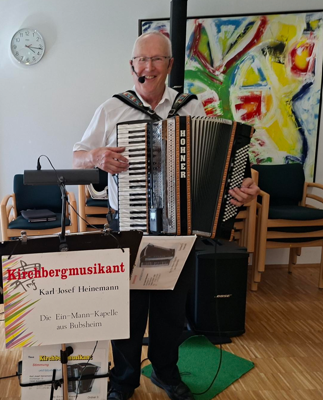 Ein älterer Mann spielt Akkordeon und trägt ein Mikrofon-Headset, während er vor einem bunten Gemälde in einem Raum mit Stühlen steht.