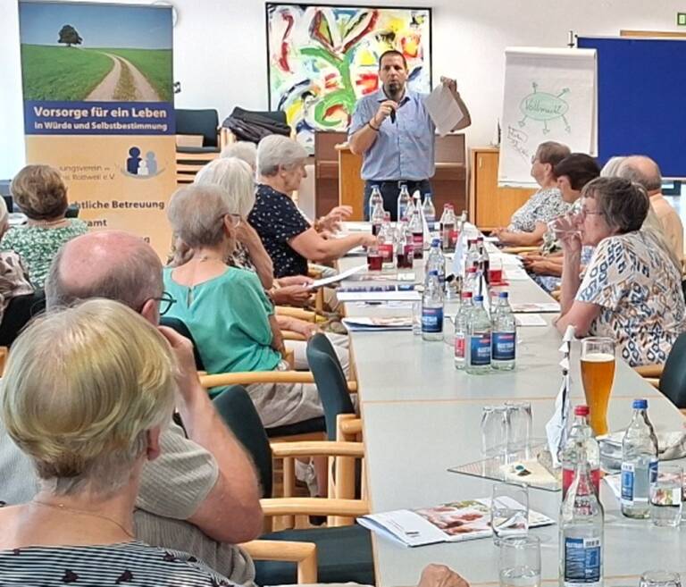 Ein Mann hält ein Mikrofon und spricht zu einer Gruppe von älteren Erwachsenen, die um einen Konferenztisch sitzen, während im Hintergrund ein Plakat mit einem Weg und einem Baum darauf zu sehen ist.