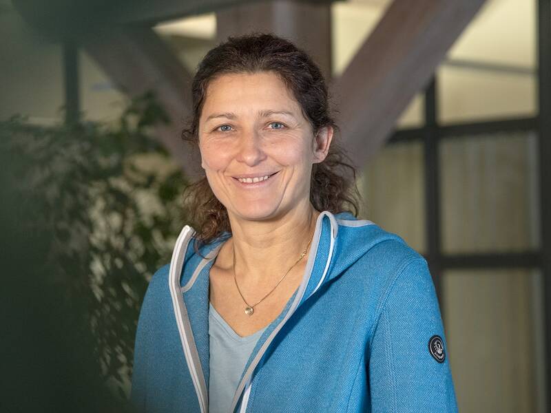 Eine lächelnde Frau in einem blauen Hoodie steht vor einem verschwommenen Hintergrund aus Pflanzen und einem Fenster.
