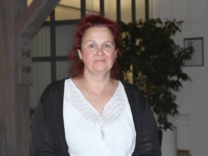Eine Frau mit roten Haaren trägt eine weiße Bluse und steht vor einem verschwommenen Hintergrund mit einer Pflanze.