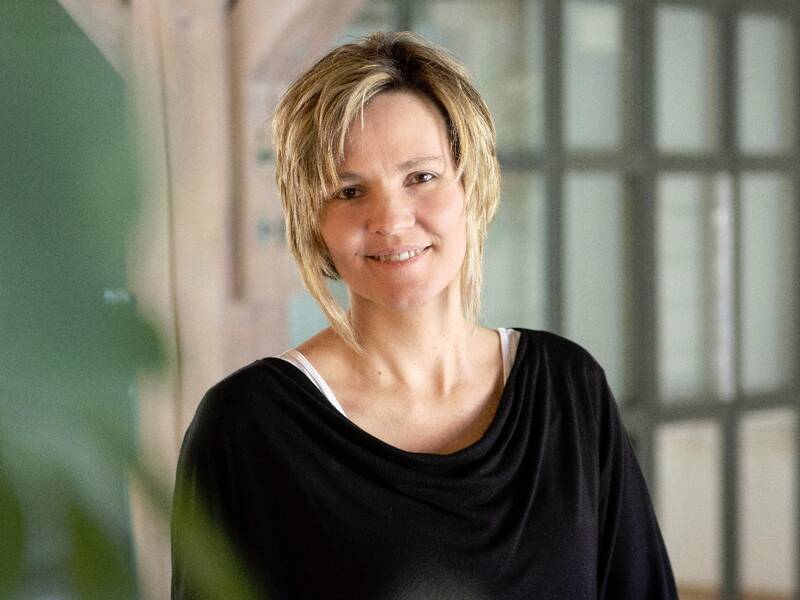 Eine Frau mit blondem Haar, die ein schwarzes Oberteil trägt, lächelt in die Kamera; im Hintergrund sind ein Fenster und Holzbalken zu sehen.