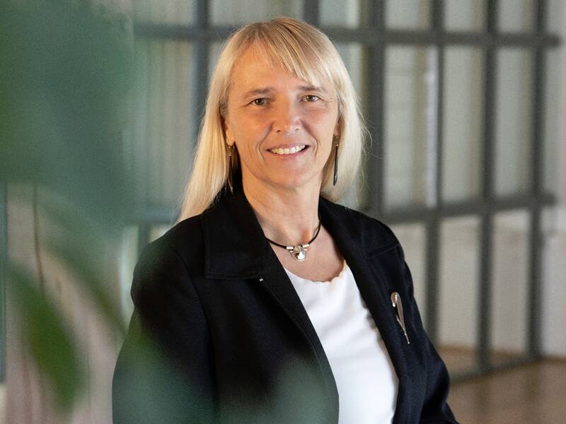 Eine Frau mit langen blonden Haaren in schwarzer Jacke und weißem Oberteil lächelt vor einem unscharfen Hintergrund mit Glasfenstern in die Kamera.