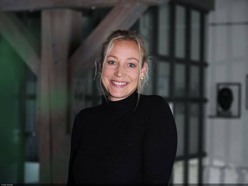 Eine lächelnde Frau mit blonden Haaren trägt ein schwarzes Oberteil, im Hintergrund ist eine Glaswand und Holzbalken zu sehen.
