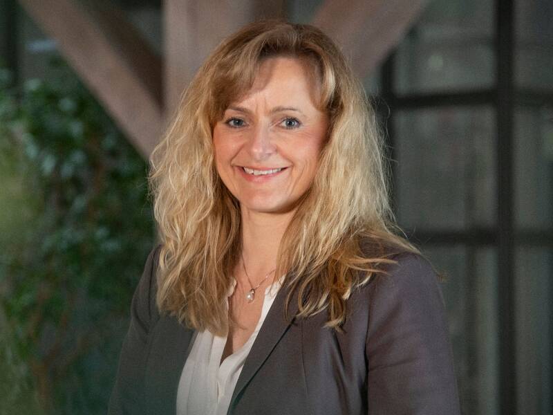 Eine Frau mit langen blonden Haaren in grauer Jacke und weißem Oberteil lächelt vor einem unscharfen Hintergrund mit Glaswand in die Kamera.
