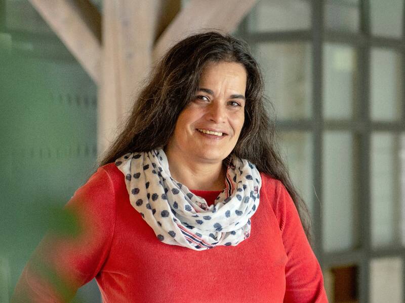 Eine Frau mit langen, dunklem Haar, die einen roten Pullover mit Schal trägt, lächelt in die Kamera und steht vor einem verschwommenen Hintergrund mit einer Glaswand und Holzbalken.