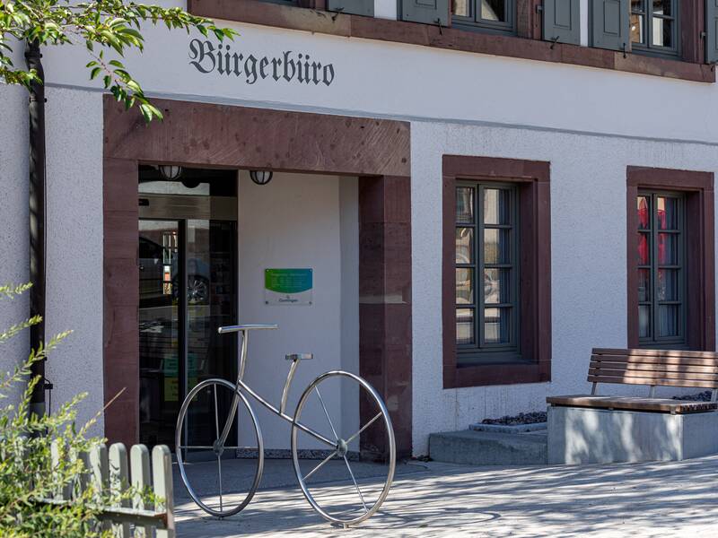 Vor einem schlichten Gebäude mit der Aufschrift "Bürgerbüro" steht eine Skulptur in Form eines großen Fahrrads, neben einer Holzbank.