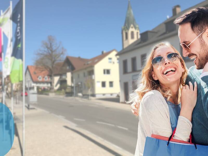 Ein lachendes Paar in Freizeitkleidung steht nah beieinander in einer sonnigen Straße mit Fahnen und Gebäuden im Hintergrund.
