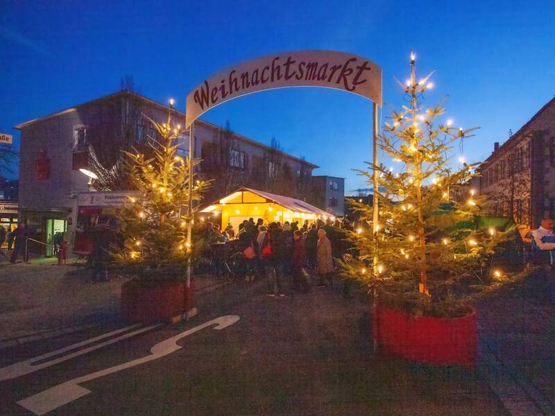 Der Weihnachtsmarkt ist festlich dekoriert mit Lichtern, geschmückten Tannenbäumen und einem großen beleuchteten Eingangsschild, während Menschen durch die Stände bummeln.