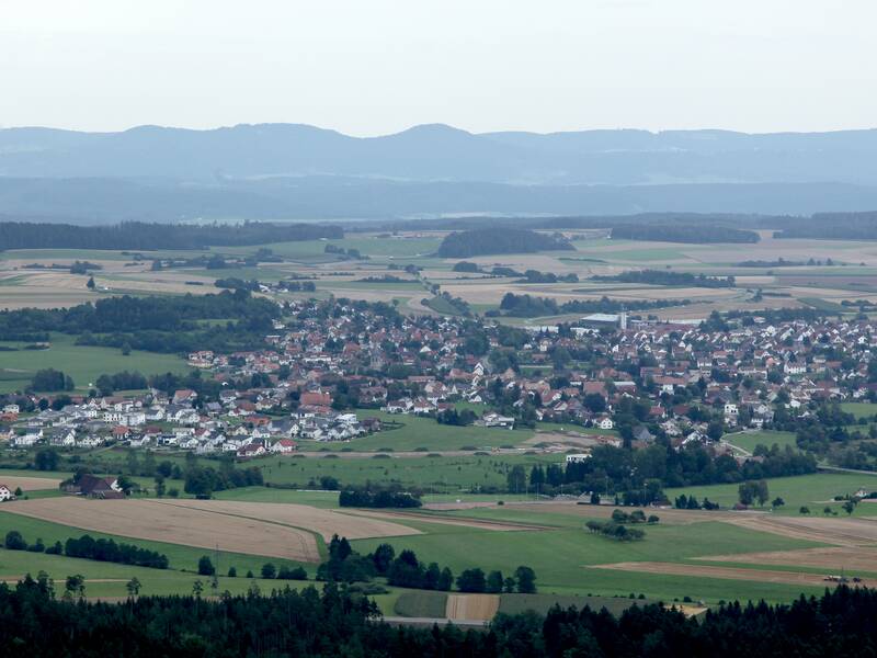 Die weite Landschaft zeigt ein Dorf umgeben von Feldern und Wäldern, mit sanften Hügeln im Hintergrund unter einem bewölkten Himmel.