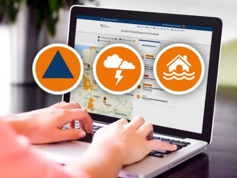 Eine Person tippt auf einem Laptop, der eine Webseite mit aktuellen Wetterwarnungen in Deutschland zeigt, daneben sind drei Symbole für Katastrophenschutz, Unwetter und Hochwasser abgebildet.