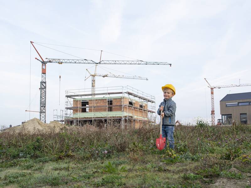 Ein Kind mit einem gelben Bauhelm und einer roten Schaufel steht vor einem im Bau befindlichen Gebäude mit mehreren Kränen im Hintergrund.