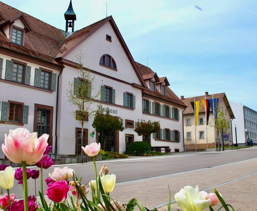 Eine Straße mit bunten Blumen im Vordergrund führt zum großen, traditionellen Dunninger Rathaus, flankiert von Fahnen an der Seite.