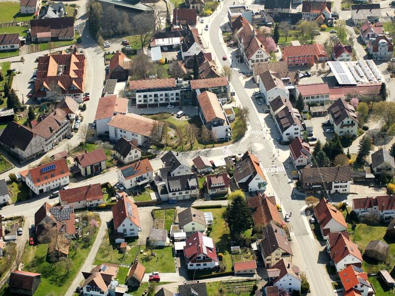 Luftaufnahme einer kleinen Stadt mit vielen dicht stehenden Häusern mit roten Dächern, grünen Gärten und mehreren Straßen, die durch das Wohngebiet führen.